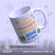 Чашка найкращій доні подарунокʼ Біла чашка з кольоровим принтом, на якому написано 'Ця чашка належить найкращій доні в світі', зображена під кутом. Ручка гладка, край рівний, фон ніжно-фіолетовий з елементами жовтих зірочок, квітів та подарунків. Чашка має глянцеву поверхню.