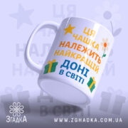 Чашка найкращій доні подарунокʼ Біла чашка з кольоровим принтом, на якому написано 'Ця чашка належить найкращій доні в світі', зображена під кутом. Ручка гладка, край рівний, фон ніжно-фіолетовий з елементами жовтих зірочок, квітів та подарунків. Чашка має глянцеву поверхню.