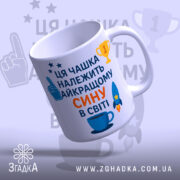 Чашка найкращому сину подарунок. Чашка білого кольору з текстом синього та оранжевого кольорів, зображенням ракети та чашки. Ручка круглої форми, край рівний, поверхня глянцева. Чашка нахилена, фон світло-синій з елементами зірок та трофеїв.