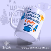 Чашка найкращому сину подарунок. Чашка білого кольору з текстом синього та оранжевого кольорів, зображенням ракети та чашки. Ручка круглої форми, край рівний, поверхня глянцева. Чашка розташована під кутом, фон світло-синій з елементами зірок та трофеїв.