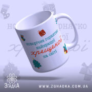 Чашка новорічна для хрещеної, біла з кольоровим принтом, зображена під кутом, ручка з округленими краями, глянець на поверхні, текст на чашці: 'новорічне горнятко найкращої хрещеної на світі', святкові малюнки навколо.