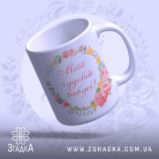 Чашка подарунок для бабусі, біла з квітковим принтом, ракурс знизу, фон світло-фіолетовий, напис червоним шрифтом "Мій чудовий бабусі!", ручка округлої форми, край гладкий, глянцевий.