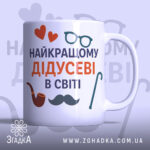 Чашка подарунок для дідуся, біла, з написом 'НАЙКРАЩОМУ ДІДУСЕВІ В СВІТІ', червоні сердечка, чорна борода, коричнева трубка, зелена тростина, ручка з класичним дизайном, гладкий край, глянцева поверхня, фон світло-фіолетовий.