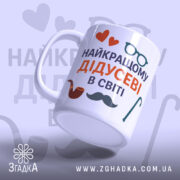 Чашка подарунок для дідуся, біла, з написом 'НАЙКРАЩОМУ ДІДУСЕВІ В СВІТІ', червоні сердечка, чорна борода, коричнева трубка, зелена тростина, ручка з класичним дизайном, гладкий край, глянцева поверхня, фон світло-фіолетовий.