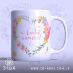 Чашка подарунок для мами, біла з квітковим принтом, ракурс з правого боку, фон світло-фіолетовий, напис 'Любій матусі!' червоним шрифтом, ручка гладка, край рівний, глянцева поверхня.