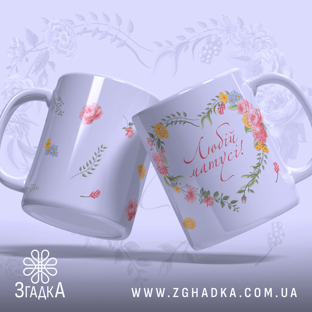 Чашка подарунок для мами, дві чашки в ракурсі, біла з квітковим принтом, фон світло-фіолетовий, напис 'Любій матусі!' червоним шрифтом, ручки гладкі, краї рівні, глянцева поверхня.