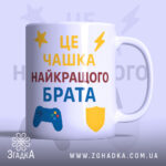 Чашка подарунок улюбленому брату. Чашка білого кольору, з текстом на передній частині, що говорить 'ЦЕ ЧАШКА НАЙКРАЩОГО БРАТА'. Принт виконаний у жовтому, червоному та синьому кольорах, з елементами у вигляді ігрового контролера та щита. Ручка стандартної форми, край гладкий, поверхня глянцева, фон світло-фіолетовий.