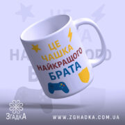 Чашка подарунок улюбленому брату. Чашка білого кольору, з текстом 'ЦЕ ЧАШКА НАЙКРАЩОГО БРАТА', розташованим на передній частині. Принт виконаний у жовтому, червоному та синьому кольорах, з елементами у вигляді ігрового контролера та щита. Ручка стандартної форми, край гладкий, поверхня глянцева, фон світло-фіолетовий.