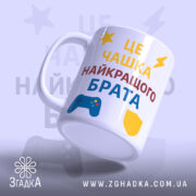 Чашка подарунок улюбленому брату. Чашка білого кольору, з текстом 'ЦЕ ЧАШКА НАЙКРАЩОГО БРАТА', розташованим на передній частині. Принт виконаний у жовтому, червоному та синьому кольорах, з елементами у вигляді ігрового контролера та щита. Ручка стандартної форми, край гладкий, поверхня глянцева, фон світло-фіолетовий.