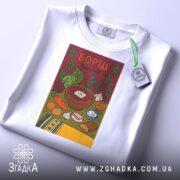 Футболка борщ українська кулінарія, біла, складена на столі, фон світло-сірий, принт з малюнком борщу та інгредієнтів, на ярлику зелена стрічка.