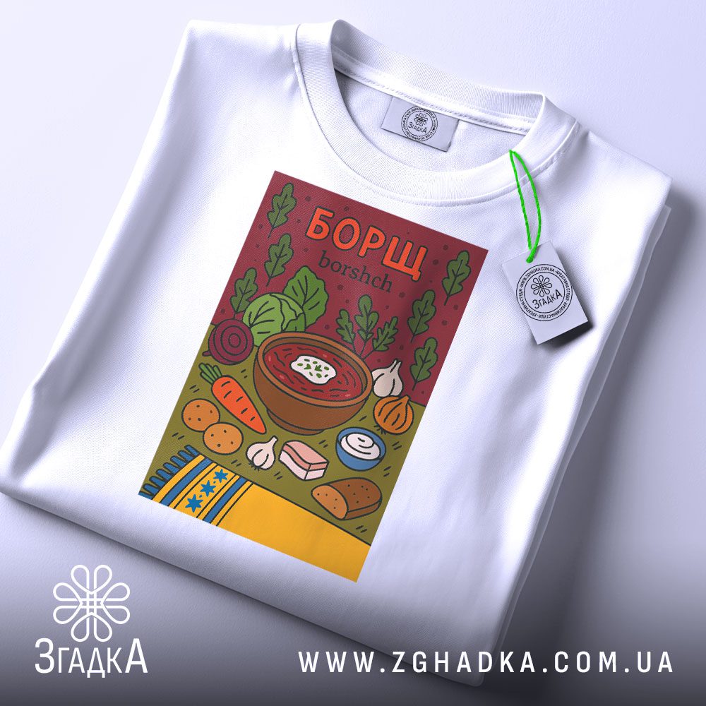 Футболка борщ українська кулінарія, біла, складена на столі, фон світло-сірий, принт з малюнком борщу та інгредієнтів, на ярлику зелена стрічка.