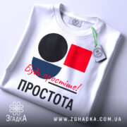 Футболка Будь простіше Простота, біла футболка, складена на столі, з принтом у вигляді кольорових геометричних фігур: чорного кола, червоного квадрата та синього півкола, текст червоним шрифтом 'Будь простіше!' та чорним 'ПРОСТОТА', на футболці висять етикетки.