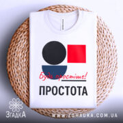 Футболка Будь простіше Простота, біла футболка, складена на плетеній кошику, з принтом у вигляді кольорових геометричних фігур: чорного кола, червоного квадрата та синього півкола, текст червоним шрифтом 'Будь простіше!' та чорним 'ПРОСТОТА', фон білий.