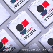 Футболка Будь простіше Простота, кілька білих футболок, складених одна на одну, з принтом у вигляді кольорових геометричних фігур: чорного кола, червоного квадрата та синього півкола, текст червоним шрифтом 'Будь простіше!' та чорним 'ПРОСТОТА', фон сірий.