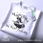 Футболка чарівна на всю голову, білого кольору, зображена в складеному вигляді, фон світло-сірий, принт з відьмою на мітлі та зірками, з зеленим етикеткою.