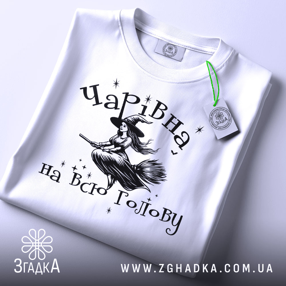 Футболка чарівна на всю голову, білого кольору, зображена в складеному вигляді, фон світло-сірий, принт з відьмою на мітлі та зірками, з зеленим етикеткою.