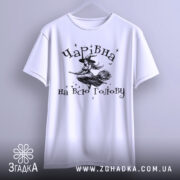 Футболка чарівна на всю голову, білого кольору, підвішена на вішалці, фон світло-сірий, принт з відьмою на мітлі та зірками.