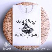 Футболка чарівна на всю голову, білого кольору, складена на плетеній кошику, фон білий, принт з відьмою на мітлі та зірками.