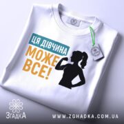 Футболка для підтримки дівчат, білого кольору, зображена на столі, фон світло-сірий, принт містить текст "ЦЯ ДІВЧИНА МОЖЕ ВСЕ!" у жовтому та блакитному кольорах, силует дівчини з піднятою рукою, на ярлику є зелена стрічка.