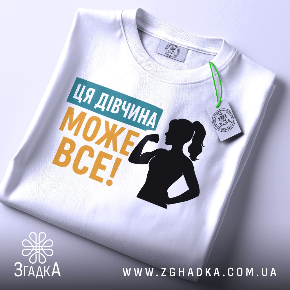 Футболка для підтримки дівчат, білого кольору, зображена на столі, фон світло-сірий, принт містить текст "ЦЯ ДІВЧИНА МОЖЕ ВСЕ!" у жовтому та блакитному кольорах, силует дівчини з піднятою рукою, на ярлику є зелена стрічка.