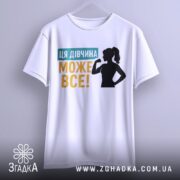 Футболка для підтримки дівчат, білого кольору, підвішена на вішалці, фон світло-сірий, принт містить текст "ЦЯ ДІВЧИНА МОЖЕ ВСЕ!" у жовтому та блакитному кольорах, силует дівчини з піднятою рукою.