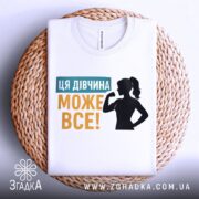 Футболка для підтримки дівчат, білого кольору, зображена на плетеному кошику, фон світло-сірий, принт містить текст "ЦЯ ДІВЧИНА МОЖЕ ВСЕ!" у жовтому та блакитному кольорах, силует дівчини з піднятою рукою.