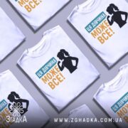 Футболка для підтримки дівчат, білого кольору, зображена на столі, фон світло-сірий, кілька футболок складені одна на одну, принт містить текст "ЦЯ ДІВЧИНА МОЖЕ ВСЕ!" у жовтому та блакитному кольорах, силует дівчини з піднятою рукою.