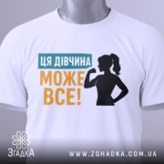 Футболка для підтримки дівчат, білого кольору, зображена на манекені, фон світло-сірий, принт містить текст "ЦЯ ДІВЧИНА МОЖЕ ВСЕ!" у жовтому та блакитному кольорах, силует дівчини з піднятою рукою.