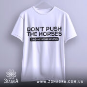 Футболка don't push the horses, біла, підвішена на вішалці, фон світло-сірий, принт чорного кольору з текстом "DON'T PUSH THE HORSES" та українським перекладом "НЕ ЖЕНИ КОНЕЙ".