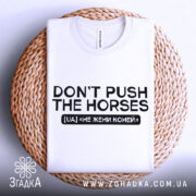 Футболка don't push the horses, біла, складена на кошику, фон світло-сірий, принт чорного кольору з текстом "DON'T PUSH THE HORSES" та українським перекладом "НЕ ЖЕНИ КОНЕЙ".