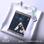 Футболка дрони не сплять, біла, розкладена на поверхні, фон світло-сірий, принт з дронами, українським гербом та силуетом людини, на ярлику зелена стрічка.