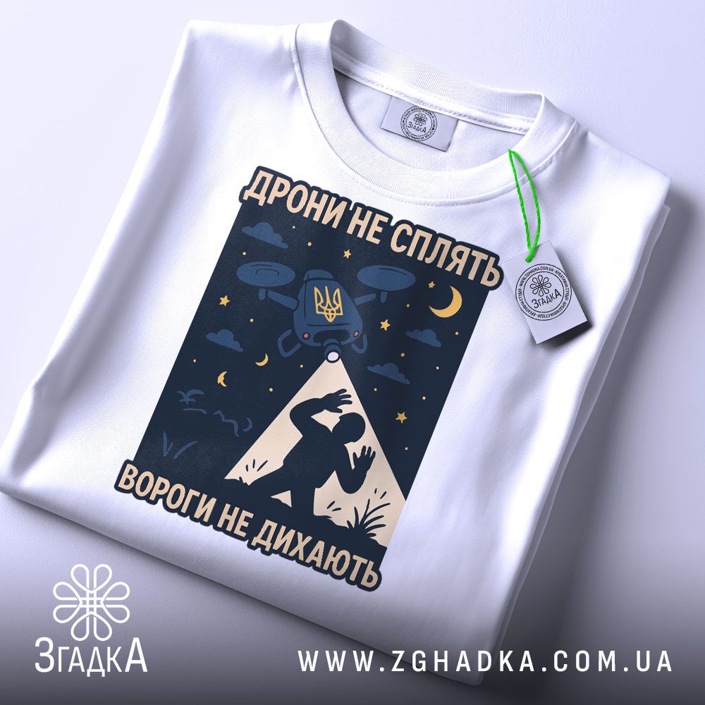 Футболка дрони не сплять, біла, розкладена на поверхні, фон світло-сірий, принт з дронами, українським гербом та силуетом людини, на ярлику зелена стрічка.