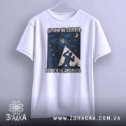 Футболка дрони не сплять, біла, підвішена на вішалці, фон світло-сірий, принт з дронами, українським гербом та силуетом людини, деталі принта чітко видимі.