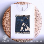 Футболка дрони не сплять, біла, складена на плетеній кошику, фон світло-сірий, принт з дронами, українським гербом та силуетом людини, видно етикетку.