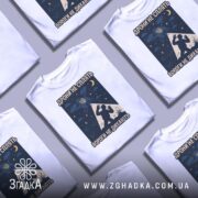 Футболка дрони не сплять, біла, складена на поверхні, фон світло-сірий, принт з дронами, українським гербом та силуетом людини, кілька футболок розкладені поруч.