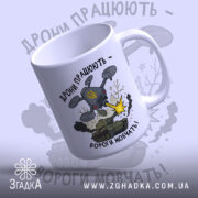 Футболка дрони працюють подарунок. Чашка білого кольору з глянцевою поверхнею, зображення дрону та танка на передній частині. Принт містить напис 'Дрони працюють - вороги мовчать!' та елементи графіки, такі як вибухи та дим. Ручка чашки класична, з округлою формою, край чашки гладкий.
