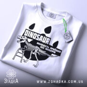 Футболка динозавр без фільтрів, біла, складена на плоскій поверхні, фон світло-сірий, принт з чорним малюнком динозавра на драбині та текстом, на комірці видна етикетка.