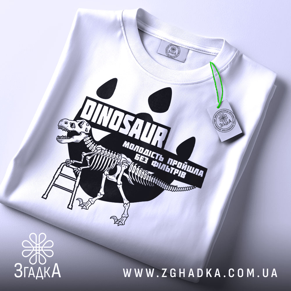 Футболка динозавр без фільтрів, біла, складена на плоскій поверхні, фон світло-сірий, принт з чорним малюнком динозавра на драбині та текстом, на комірці видна етикетка.