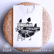 Футболка динозавр без фільтрів, біла, складена на плоскій поверхні з плетеного матеріалу, фон світло-сірий, принт з чорним малюнком динозавра на драбині та текстом.