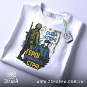 Футболка герої тримають стрій. Біла футболка, на якій зображено той же принт, але з видимим етикеткою на комірці. Принт яскравий, з акцентом на військову тематику та патріотичний зміст.