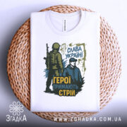 Футболка герої тримають стрій. Біла футболка, складена на плетеній основі, з добре видимим принтом. Принт містить патріотичний напис та зображення двох чоловіків.