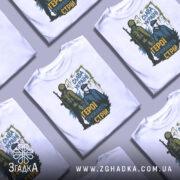 Футболка герої тримають стрій. Біла футболка, розкладена на фоні сірого кольору, з кількома складеними футболками поруч. Принт яскравий, з акцентом на патріотичну тематику.