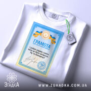 Футболка грамота з написом, білого кольору, зображена на столі, фон світло-сірий, принт з грамотою у блакитних та жовтих кольорах, текст на українській мові, на футболці є етикетка з зеленим шнурком.