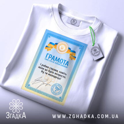Футболка грамота з написом, білого кольору, зображена на столі, фон світло-сірий, принт з грамотою у блакитних та жовтих кольорах, текст на українській мові, на футболці є етикетка з зеленим шнурком.