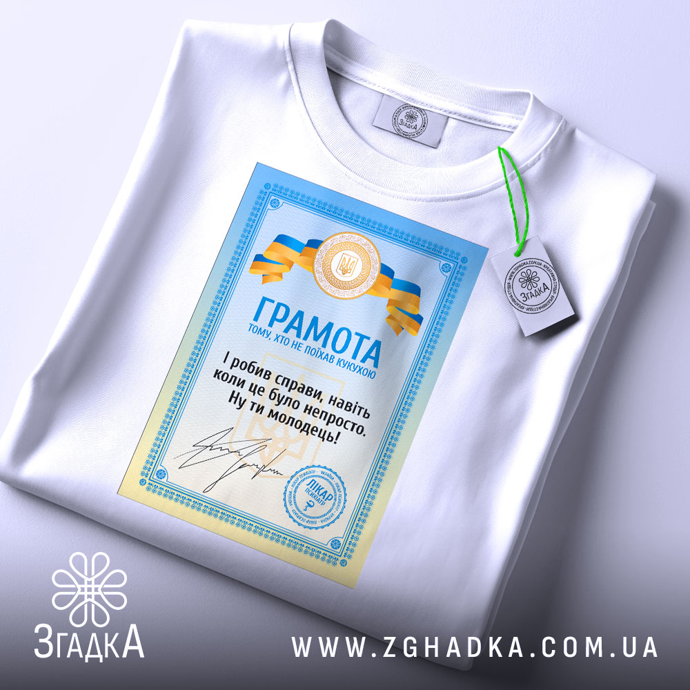 Футболка грамота з написом, білого кольору, зображена на столі, фон світло-сірий, принт з грамотою у блакитних та жовтих кольорах, текст на українській мові, на футболці є етикетка з зеленим шнурком.
