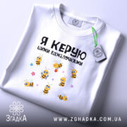 Футболка я керую бджілочками, білого кольору, складена на поверхні, фон світло-сірий, принт з бджілками та написом, на футболці є етикетка.