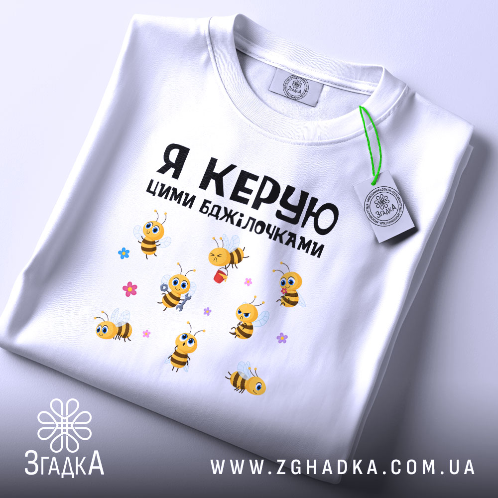 Футболка я керую бджілочками, білого кольору, складена на поверхні, фон світло-сірий, принт з бджілками та написом, на футболці є етикетка.