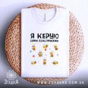 Футболка я керую бджілочками, білого кольору, складена на плетеній основі, фон світло-сірий, принт з бджілками та написом, видно деталі горловини.