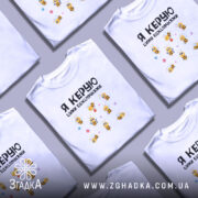 Футболка я керую бджілочками, білого кольору, складена на поверхні, фон світло-сірий, кілька футболок розташовані поруч, принт з бджілками та написом.