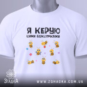 Футболка я керую бджілочками, білого кольору, зображена на манекені, фон світло-сірий, принт з бджілками та написом, видно деталі горловини та рукава.